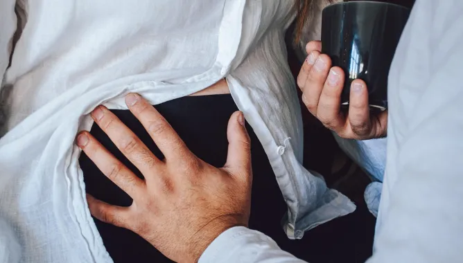 Mężczyzna głaszcze brzuch ciężarnej kobiety. W dłoni trzyma kubek z napojem.